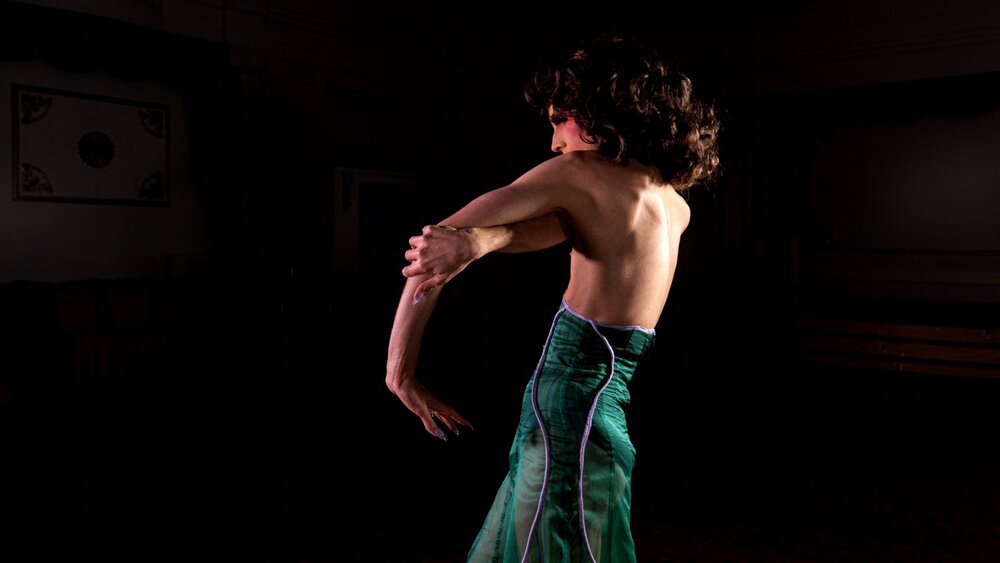 A person standing on profile. They have fair skin, pink makeup on their cheek and thick, brown curly hair that reaches just past their shoulders. They are not wearing a top, and their arms and back are muscular, light shining on their back illuminating its contours. their arms curl around each other, outstretched in front of them. one hand grips the other forearm. From their waist down they are wearing a sea green, luminous skirt with purple edging. They have matching, long sharp purple nails. The space around them is all black.