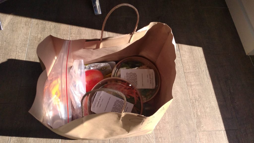 Photo of a brown paper bag in the centre of the frame. The angle is above the bag looking into the contents, which include 2 sandwich bags, two salads in take away boxes with a clear top lid and a tomato., as well as indeterminable contents. The paper bag is on a dark wooden floor lit by natural sunlight coming in from the right side off screen. Top centre of the image is the end of a silver chair leg, with a small black wheel. In the right corner a part of a white wall comes into frame.