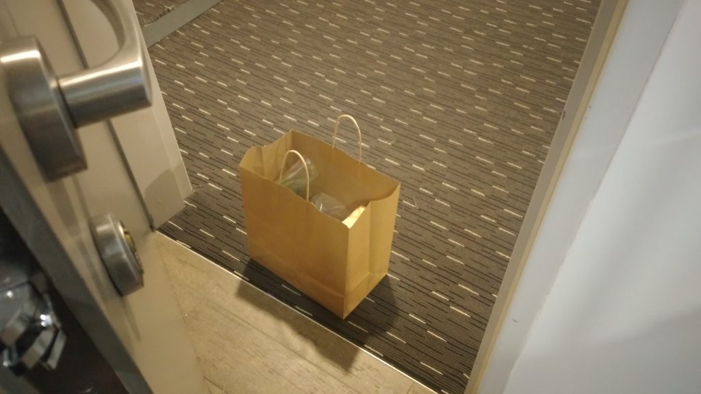 Image is a photo taken of a brown paper bag outside a hotel room door. The photo is taken from inside the room looking down at the bag in the hotel corridor. The centre of the image is the paper bag which is open and contains indeterminable contents in plastic bags. The floor is divided by the doorway, inside the room is wooden slats and the corridor is carpeted dark grey with thin sporadic white and black stripes. On the left side of is a close up of the silver door handle, and door locking mechanism and the right side is a white wall.