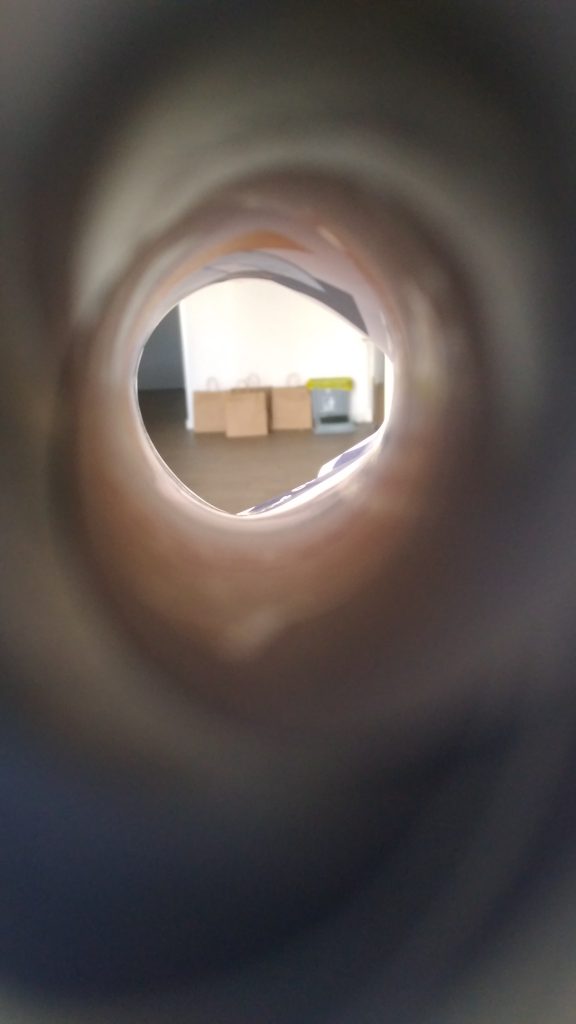 Image is of three blurry paper bags in a row, and a small rubbish bin next to them, against a white wall. The photo is taken through a small cylindrical tunnel made from a rolled up piece of paper. The tunnel creates a telescope illusion.