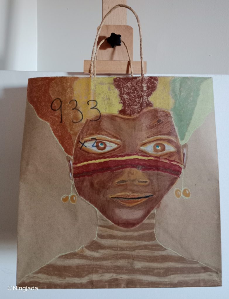 Image is a photo of a brown paper bag which has been drawn on, hanging on a wooden easel, in front of a white wall. The drawing is of a women. The woman’s face is centre of the bag, they have dark coloured skin, and brown eyes and have horizontal stripes across their cheeks and nose, which are red, yellow earthy colours. . Their hair floats upwards and is multicoloured vertical stripes of brown, yellow and green earthy colours. The woman has identical cherry shaped orange earrings. Their neck and shoulders are striped white and brown horizontally. The number “933” is written top left over the hair and partial forehead of the woman.