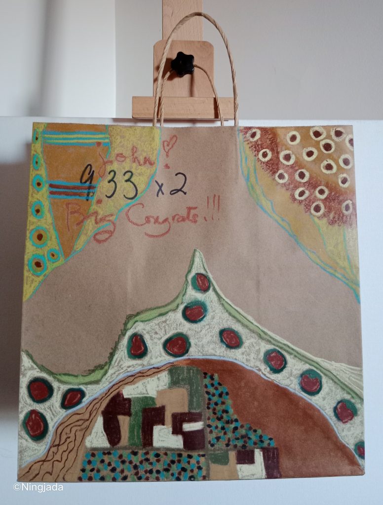 Image is a photo of a brown paper bag which has been drawn on, hanging on a wooden easel, in front of a white wall. There are three sections drawn in. The top left of the bag has triangle shapes which contain circles and stripes of light blue, dark red, yellow and orange colours. The top right of the bag is coloured in with orange and dark red shades, with white and brown centred circles. The bottom of the bag is coloured with dark brown, red, green and blue shades. It contains block like shape patterns and is boarded by dark red centred circles outlined with a dark green. “John love heart… big congrats!!!” is written in red with “933x2” written in black top left of the bag.
