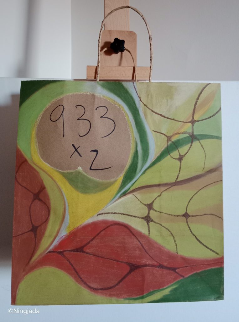 Image is a photo of a brown paper bag which has been drawn on, hanging on a wooden easel, in front of a white wall. “933x2” is written top left of the bag in black pen. The writing is encased by the drawing which is made up of leaf like shapes in red, yellow, light and dark green shades. Thin brown lines are drawn within these shapes creating a web like effect.