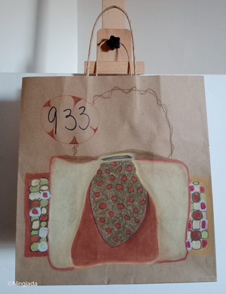 Image is a photo of a brown paper bag with a drawing on it, hanging on a wooden easel, in front of a white wall. The handbag is predominantly off white, and dark red in the centre. It has a dark grey buckle flap with red dots on it. The handle is a wiriey brown handle. Both sides of the handbag have protruding sides that have a checkered pattern of white, red circles and green squares. “933” is written in black pen and circled with a red marker and small triangles pointed at the number.