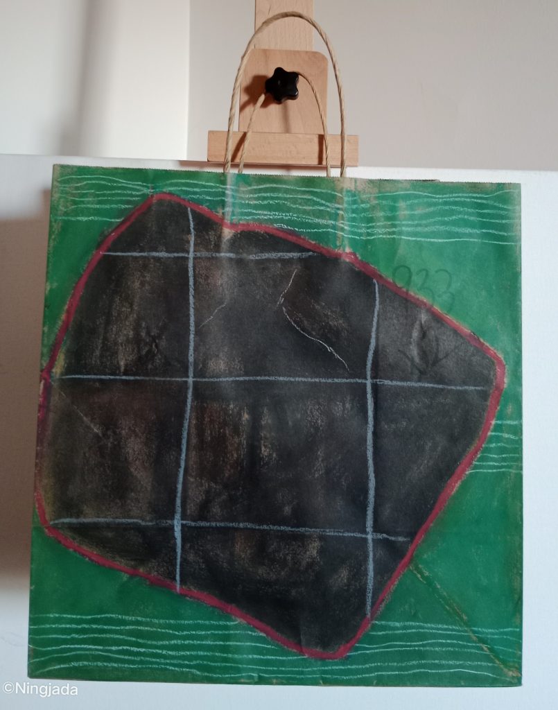 Image is a photo of a brown paper bag which has a picture drawn on it, hanging on a wooden easel, in front of a white wall. The image is a black square on a tilt, outlined in red. The square has white lines over it creating a numeric table shape. Behind the black square is a green background with horizontal white lines clumped together, top, middle and bottom of the bag. The number “933” is faded under the green background, top right of the bag.
