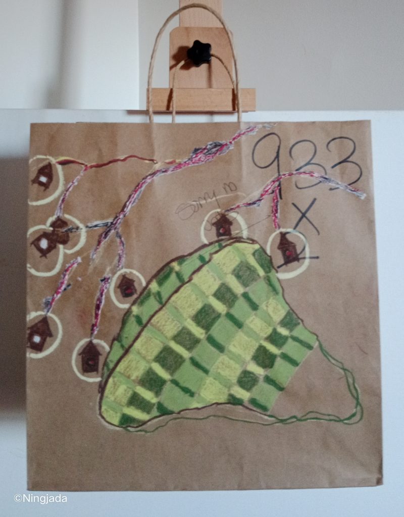 A brown paper bag has a circular shape drawn on it in the centre. The shape consists of a square and thin rectangle shaped pattern, with shades of dark green, light green and lime green. The top left of the bag has multiple small brown house shaped like figures, all with a white circle around each. Each are connected through a root like system drawn on with shades of pink, red, blue and white in it. The top right of the bag under the layers of the roots is the number “933 x” as well as “sorry no oats”. The bag is hanging on a wooden easel in front of a white wall.