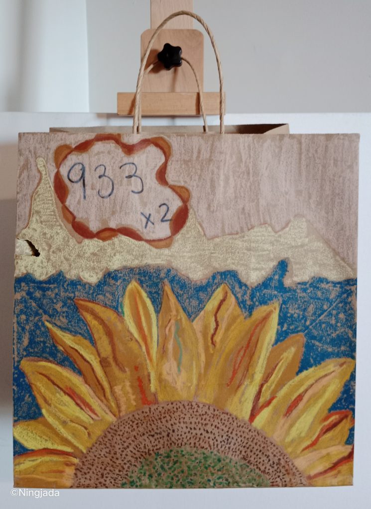 A brown paper bag has a sunflower drawn on it. The sunflower is at the bottom of the bag and half of it is cut off by the bottom of the bag. The centre of the flower is a mustard green, surrounded by a brown outer ring. The petals are a mixer of yellow, orange and red shades. Behind the flower is an ocean blue colour that continues up till just above centre of the bag. Above the blue is a pale white rocklike formation across the top of the bag. The top of the bag is coloured with a pale white. The number “933x2” is in the top left of the bag and is encased by a red outline which forms a cloud like shape.