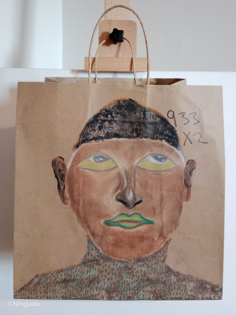 A brown paper bag has a face drawn in the centre of it. The face has light brown coloured skin, green lips outlined with a dark green, and large lime green eyes with grey pupil which are looking upwards. The person has large ears, a thin nose that is coloured dark brown. They are wearing a black beanie like head piece. The person is wearing a blue spotted skivvy. The background is not coloured in, and has “933x2” written in thin black pen, just to the right of the persons forehead. The bag is hanging on a wooden easel, in front of a white wall.