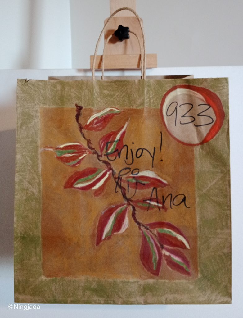 A brown paper bag has a thin vine like branch drawn in the centre with leaves attached. The leaves are a dark red, with white and green stripes on each leaf. The background of the vine is a muddy brown colour with the words “ Enjoy! {Smiley face is drawn}, Ana” layered under the vine. The vine is framed by a thick brownie green shade, around the whole outskirt of the bag. The number “933” is written on the top right and is circled in red with a white streak in the top left of the circle. The paper bag is hanging on a wooden easel in front of a white wall.