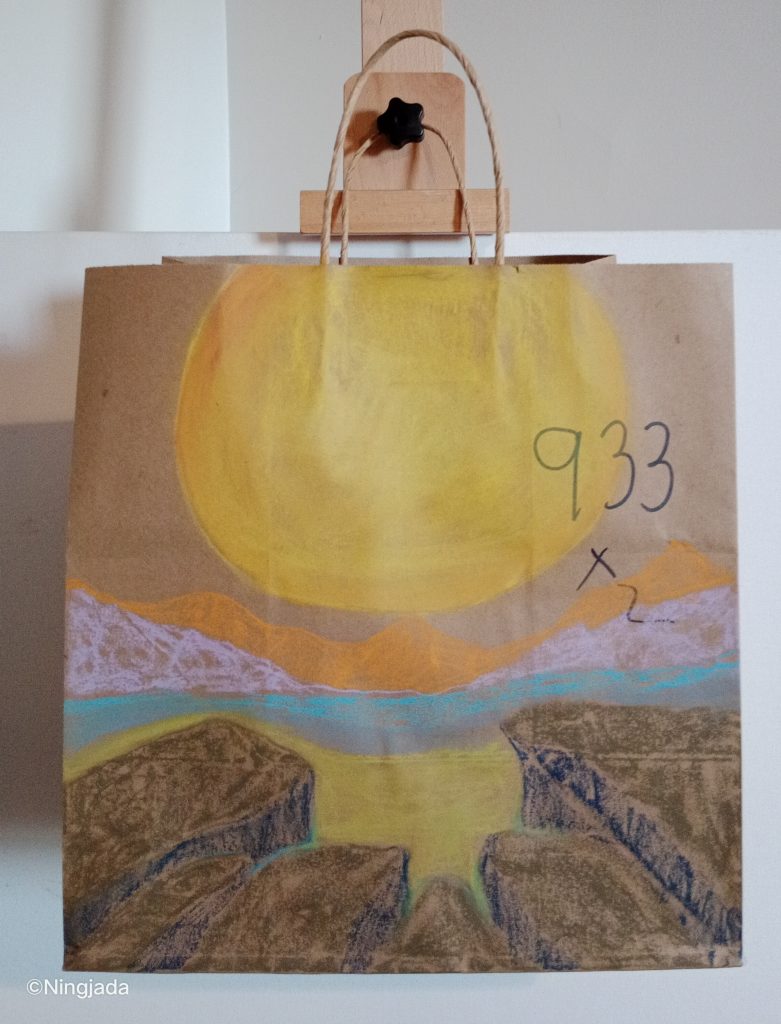 A brown paper bag has an ocean sunset drawn on it. In the top centre of the bag is a large bright yellow sun. The bottom of the bag has sea rocks pointing towards the sun, with sand in the middle above them. There is a thin horizontal line of blue ocean, above it are silhouetted mountains shaded light pink and orange. “933x2” is written in thin black pen on the right of the bag. The bag is hanging on a wooden easel, in front of a white wall.