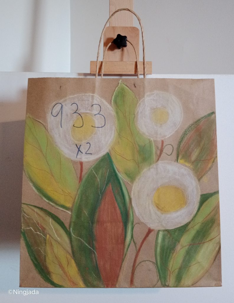 A brown paper bag has three white circular flowers drawn on it. The flowers have a yellow bud centre and a thin red stem. The flowers are surrounded by six large leaves. The leaves are a mixture of dark green, red, lime green, brown, and yellow shades. The top left flower has “933x2” thinly written in pen. The background of the bag has not been coloured in. The bag is hanging on a wooden easel, in front of a white wall.