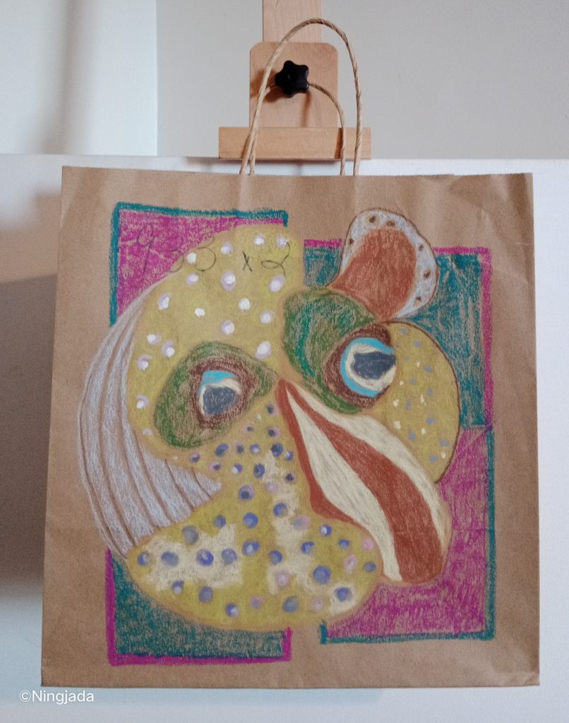 A brown bag has a obscure fishlike drawing on it. The fishlike drawing is a circle, and has two blue googly eyes in the centre of it. The shape has three different types of fins in it, the left is a striped grey fin, the right is thickly striped dark red and white, and the top right fin is rounded with dark red in the centre and grey on the outside with red dots. The fishlike shape is predominantly a mustard green with white and blue dots. Behind the fish are four squares, two of them are pink with a teal outline, and two of them are teal with a thin pink outline. The squares match diagonally. The out skirts of the bag is not coloured in. The bag is hanging on a wooden easel, in front of a white wall.
