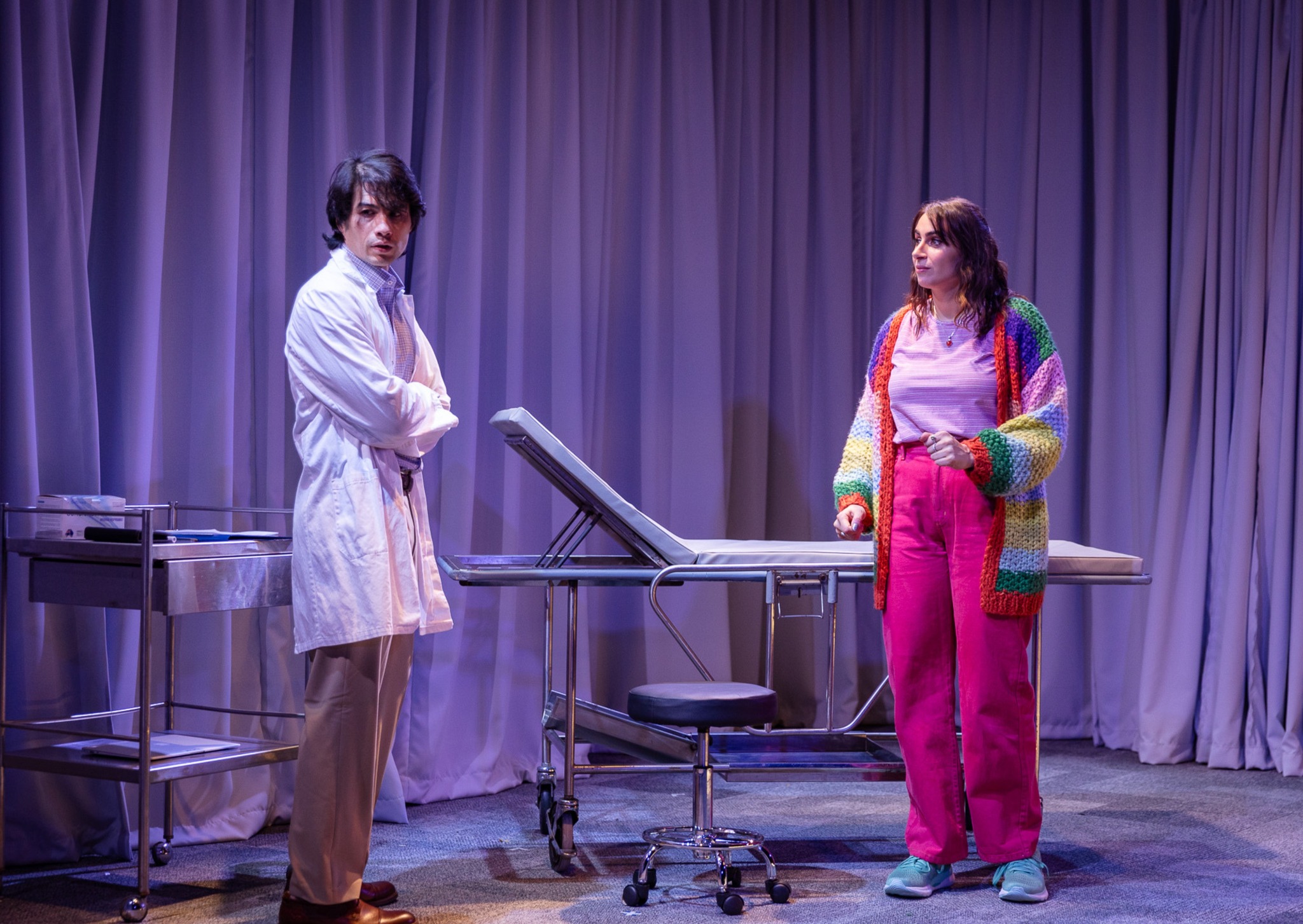 hospital room scene from telethon kid. two people stand in front of a hospital bed on wheels. One of them is a male presenting person wearing a white lab coat. He is looking away from the other with a concerned expression on his face and his arms crossed. The other person is a female presenting character with long brown hair, wearing bright pink pants and a knitted colourful jacket. She is looking at him with her eyebrows slightly raised.