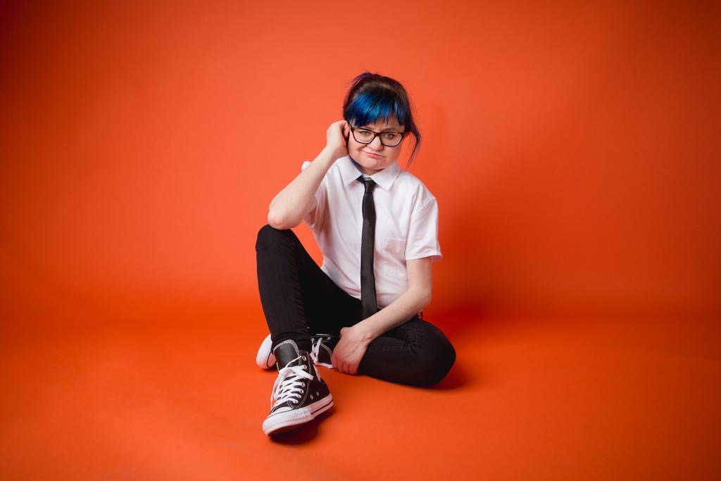 A studio photo of Jess, a fair skinned person, with dark hair except for a bright blue fringe, and black rimmed clear glasses. She sits on the ground, with one knee up, her elbow resting on it. She is wearing a white collared shirt, black tie, black tight pants and black sneakers shoes with white laces. Her mouth is in a thin line, and one eyebrow is slightly raised. The background of the photo is a deep orange, and the ground she