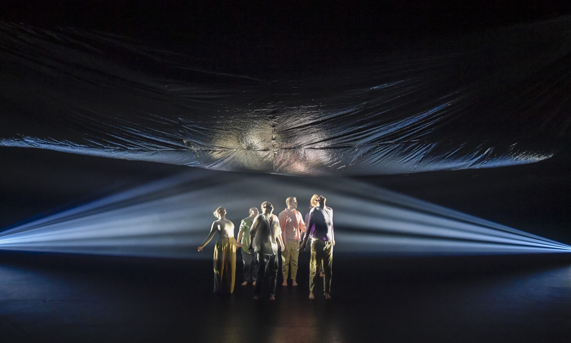 five people stand in a darkened room, wearing shirts and pants of different colours, and all looking upwards. They are gazing up at a billowing silver piece of material, that hangs above them and balloons out in the middle. Lights are shining from either side of the room onto the people and the silver material, which reflects the light off its metallic surface.